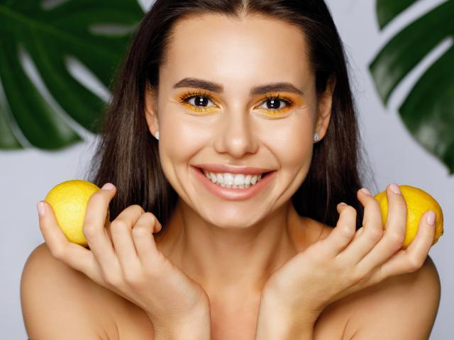 Young woman with bright yellow eyeshadow, smiling and holding lemons near face.